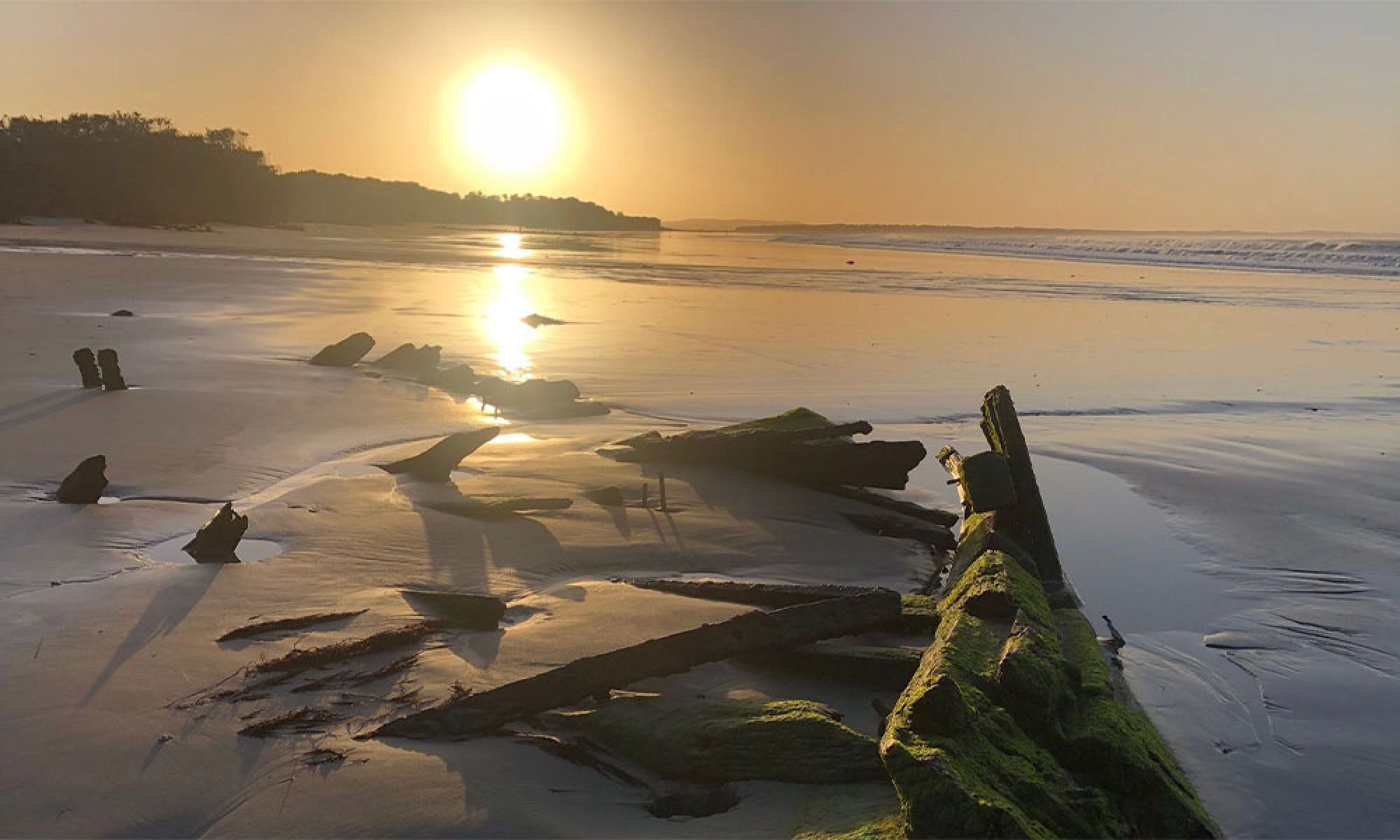 Amazon shipwreck Inverloch, washed up pieces of the ship can be seen on the beach, the wood from the ship is growing moss, The sunset can be seen behind pieces of the ship with gentle waves washing upon the sea shore. 