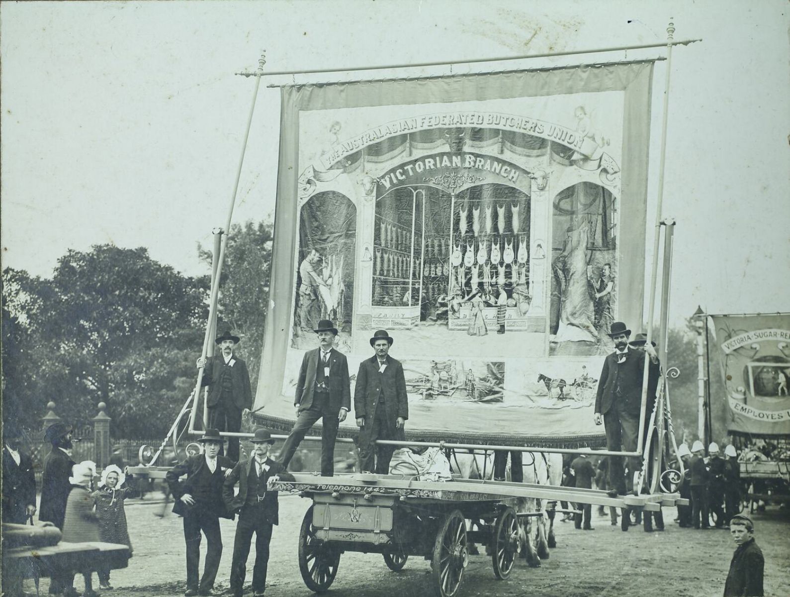 A black and white photo of large square banner on a cart with men standing at the base