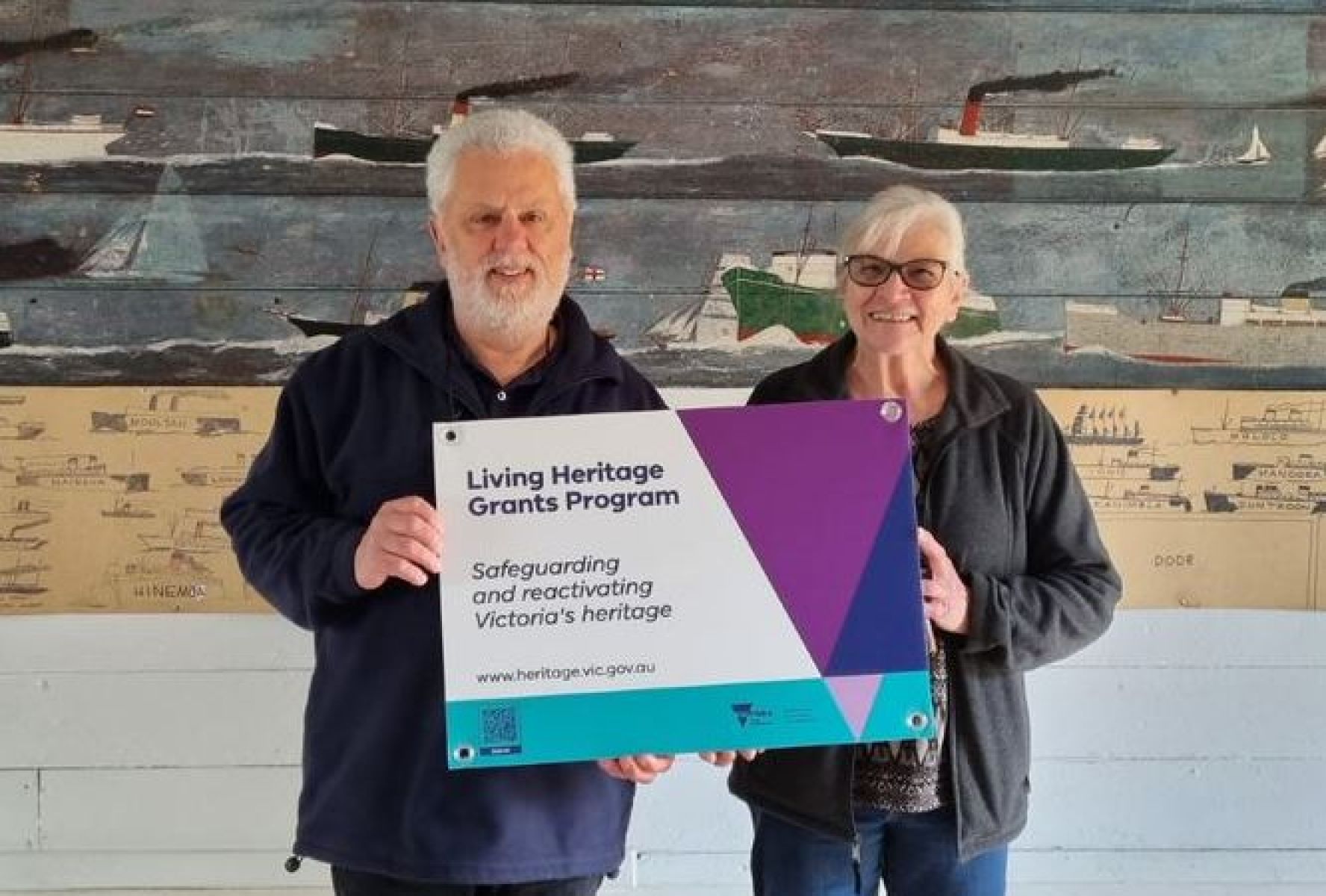 Two people hold up a sign that reads 'Living Heritage Grants Program'. They stand infront of a wall that is painted as a beach scenery with boats, sand, water and sea life. 