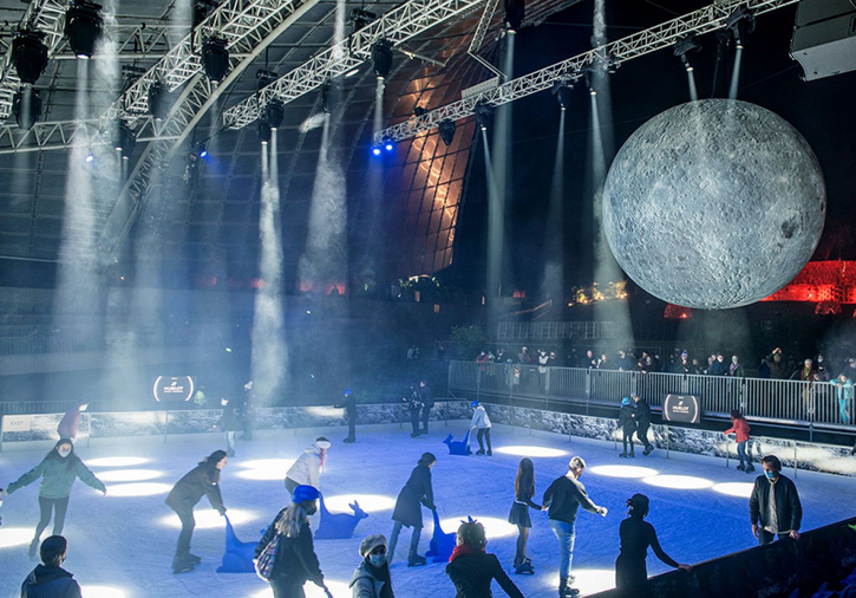 Image shows an illuminated ice ring with a full moon hovering behind it.  People skate on the ring at night time in Melbourne as part of the festival.