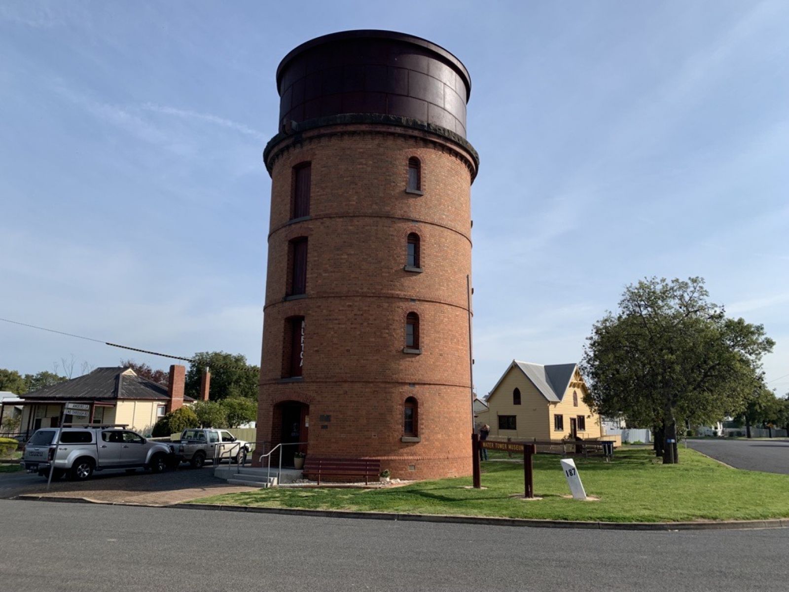 Image shows the water tower from a distance. The tower is made out of red brick and a house can be seen behind it. 