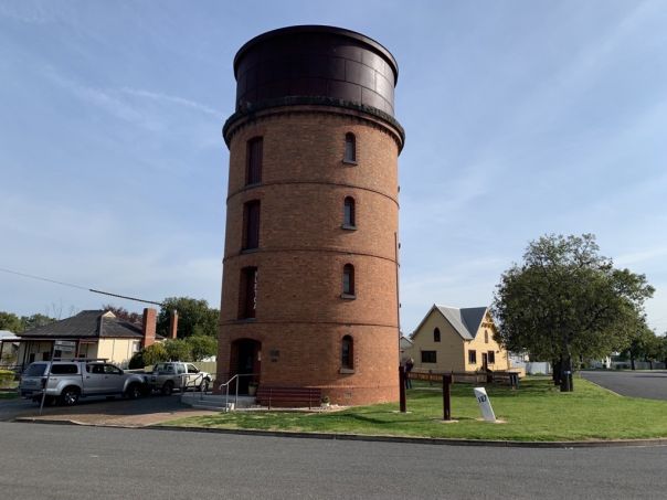Image shows the water tower from a distance. The tower is made out of red brick and a house can be seen behind it.