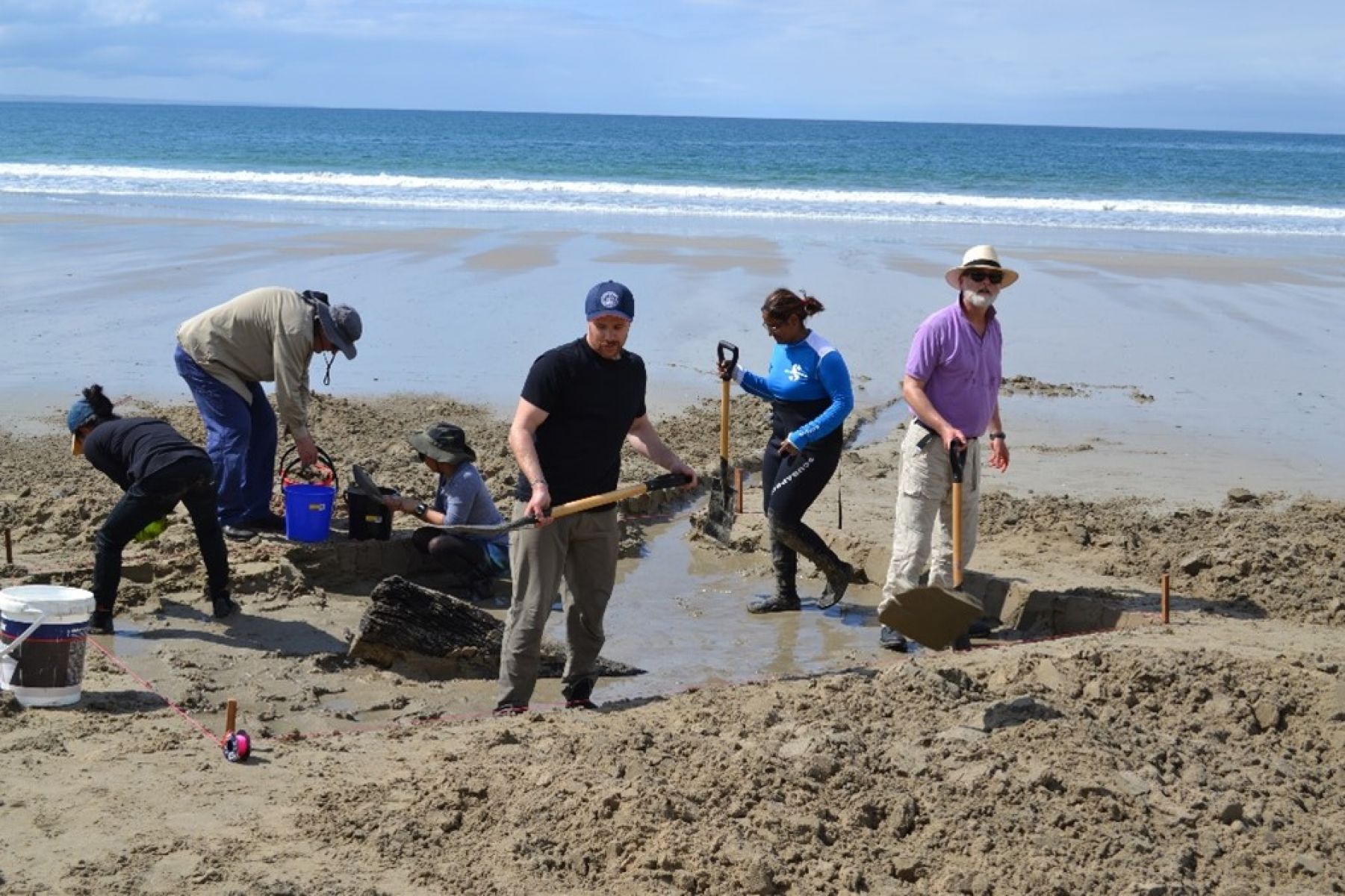 Heritage and university staff can be seen on a beach at the site of the Amazon wreck. Six people can be seen with shovels and buckets digging in the wet sand around the wreck. The ocean can be seen in the background. 