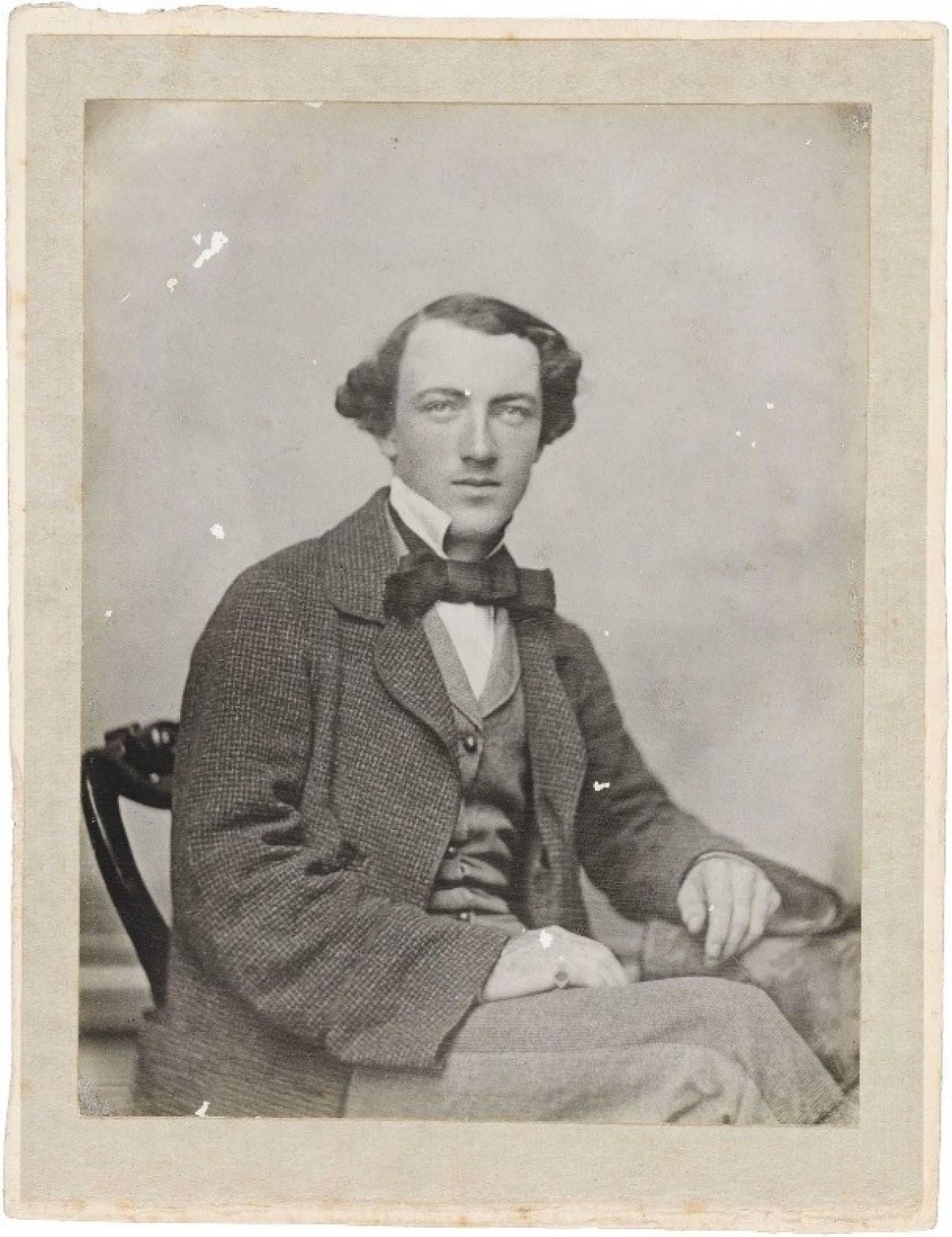 A sepia-toned photograph with some damage from age, showing a man seated on a chair, facing forward but angled slightly to his left. His left arm rests on the arm rest, his right arm rests on his knee. He is looking straight into the camera with a neutral expression and is dressed in a bow tie, jacket and matching vest with a starched white shirt collar sitting up along his jawline.
